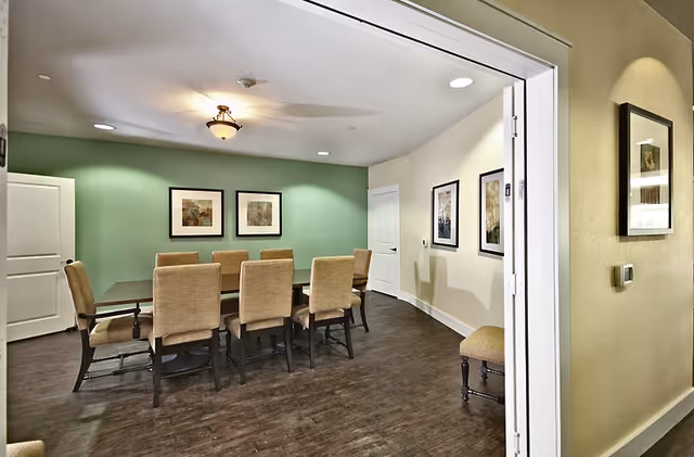A dining room with a long rectangular table surrounded by eight beige upholstered chairs. The room has a green accent wall with two framed pictures and a beige wall with two more framed pictures. The floor is dark wood, and there is a ceiling light fixture and recessed lighting.