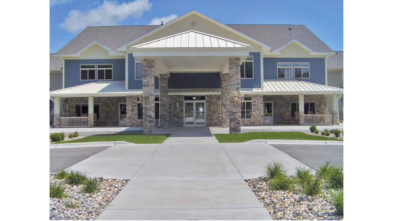 Front exterior view of a two-story retirement community building with stone pillars, a covered entrance, and a paved walkway leading to the main doors. The building has blue siding and multiple windows under a clear blue sky.