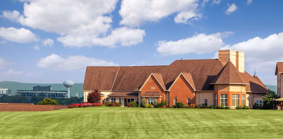 Large brick and stucco building with steep gabled roofs set behind a wide manicured lawn under a blue sky.
