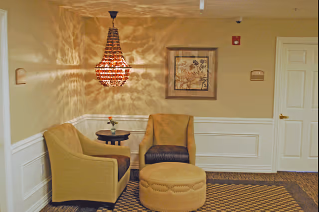 A cozy seating area in a senior living facility with two upholstered chairs, a round ottoman, a small side table with a flower vase, a decorative hanging light fixture casting patterned shadows on the wall, and a framed artwork. The walls are beige with white wainscoting, and there is a white door with a gold handle on the right.
