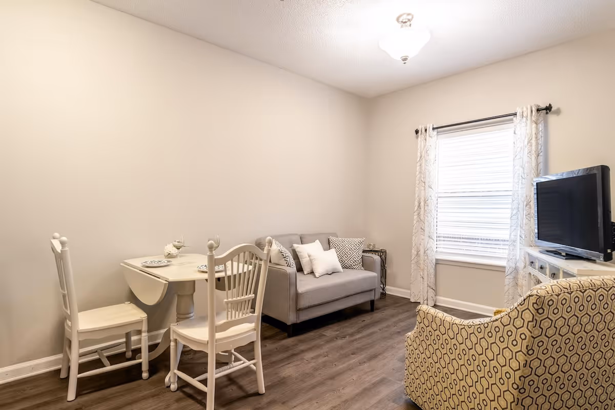A small living area with a gray loveseat adorned with white and patterned pillows, a patterned armchair, a white drop-leaf dining table with two white chairs, a window with white patterned curtains, and a flat-screen TV on a white cabinet. The room has light-colored walls and wood flooring.