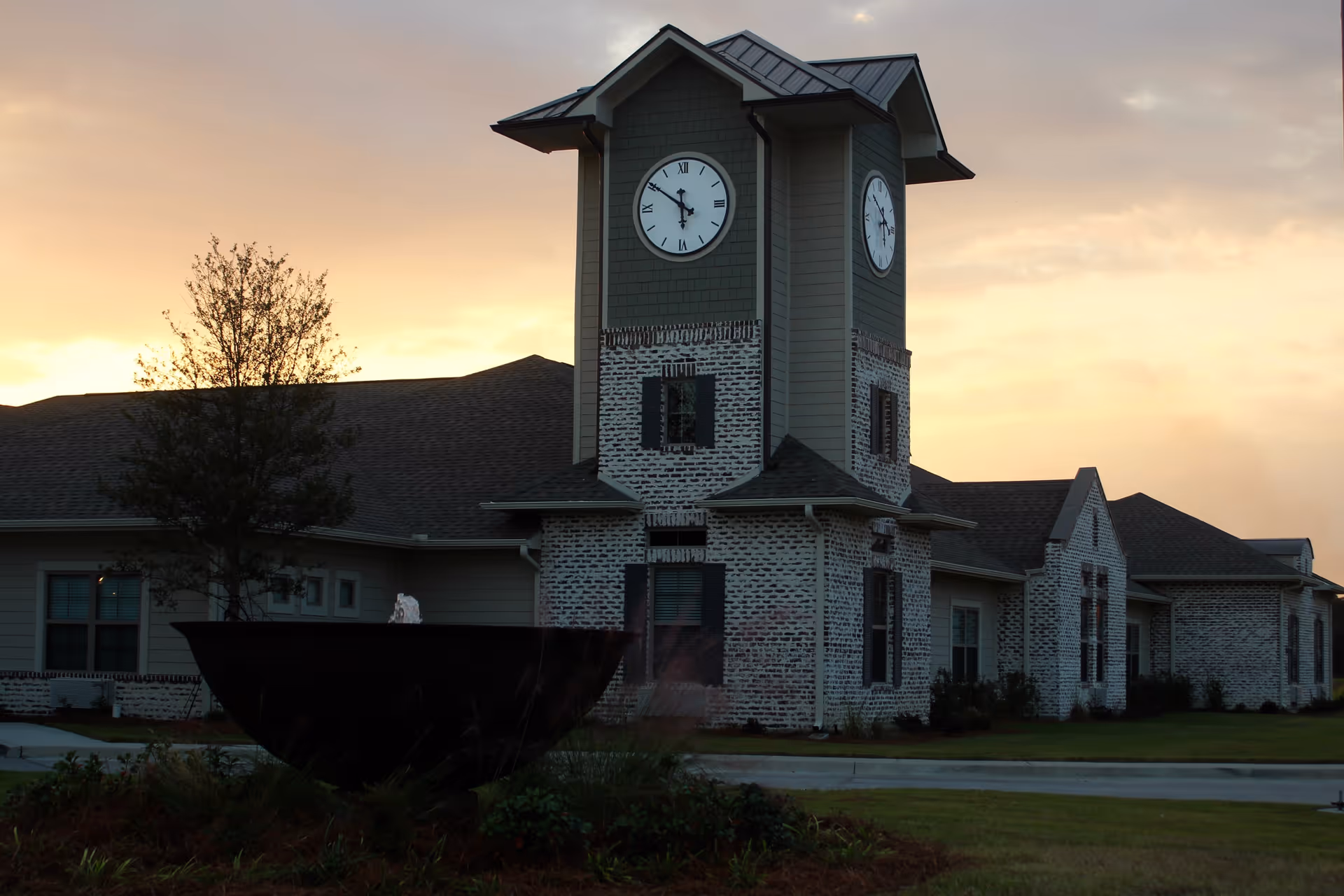 Exterior view of a senior living facility building at sunset featuring a clock tower with two visible clock faces, a large dark fountain in the foreground, and a well-maintained lawn with some shrubs and a tree.
