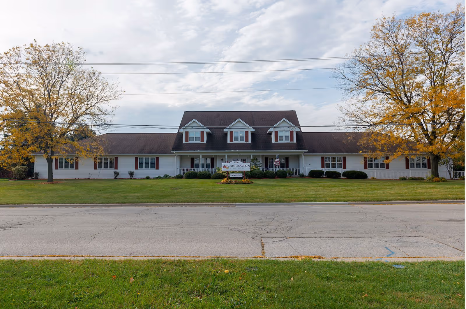 Front exterior view of a single-story senior living facility building with a dark roof, white siding, and red shutters. There are two large trees with autumn-colored leaves on either side of the building, a well-maintained lawn, and a sign in front of the entrance. The sky is partly cloudy.