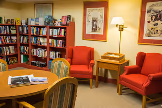 Cozy reading room with bookshelves, a round table and chairs, and two red armchairs beside a lamp and side table.