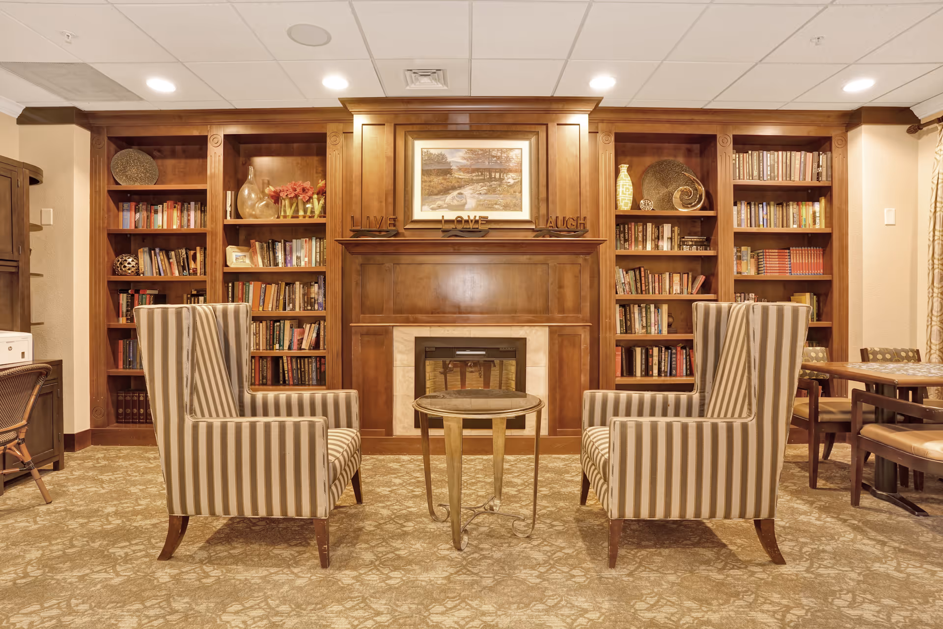 A cozy living room area with two striped armchairs facing a small round table. Behind the chairs is a wooden fireplace mantel with a framed landscape painting above it and decorative words 'LIVE LOVE LAUGH'. Flanking the fireplace are built-in wooden bookshelves filled with books and decorative items. To the right, there is a wooden table with chairs. The room has warm lighting and a patterned carpet.