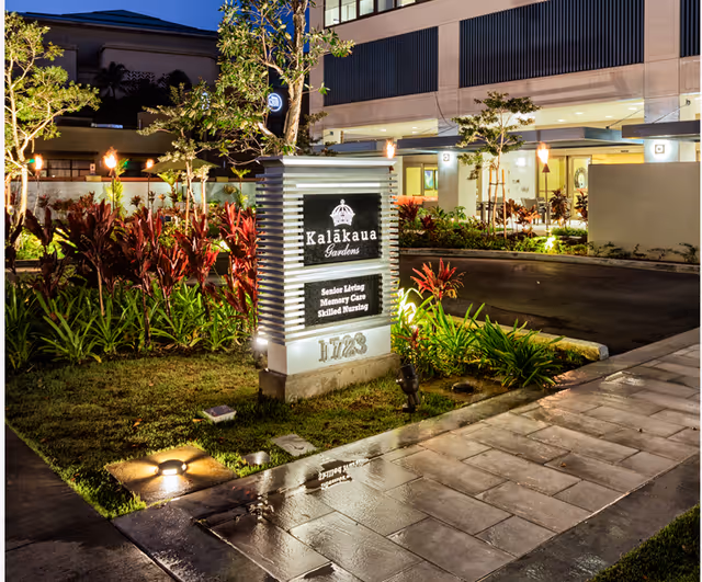 Illuminated sign for Kalākaua Gardens senior living facility at night, surrounded by landscaped plants and a paved walkway, with the building entrance visible in the background.