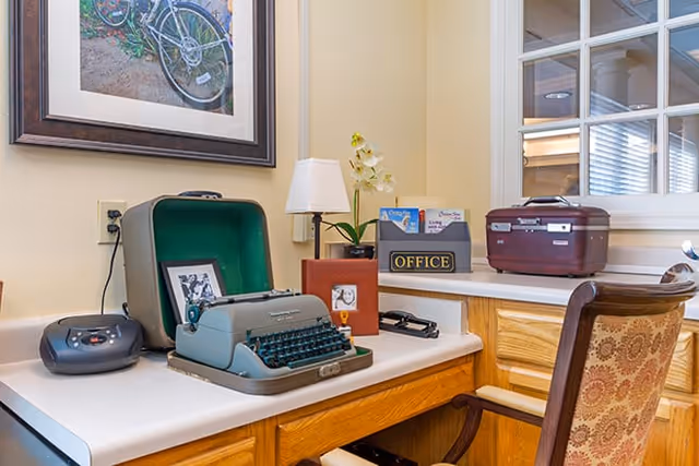 A wooden desk with a vintage typewriter in an open green case, a small lamp, a framed photo, a black office sign, and a maroon suitcase. A patterned chair is positioned in front of the desk. A framed picture of a bicycle hangs on the wall above the desk.