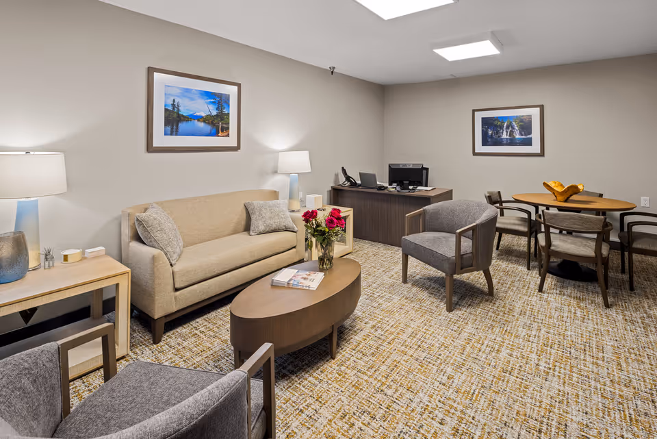 A cozy senior living community room featuring a beige sofa with two gray cushions, two gray armchairs, a wooden oval coffee table with a vase of red roses and magazines, a wooden side table with a lamp and decorative items, a wooden desk with a computer and phone, and a round dining table with four chairs and a decorative centerpiece. The walls are light beige with two framed landscape photographs.