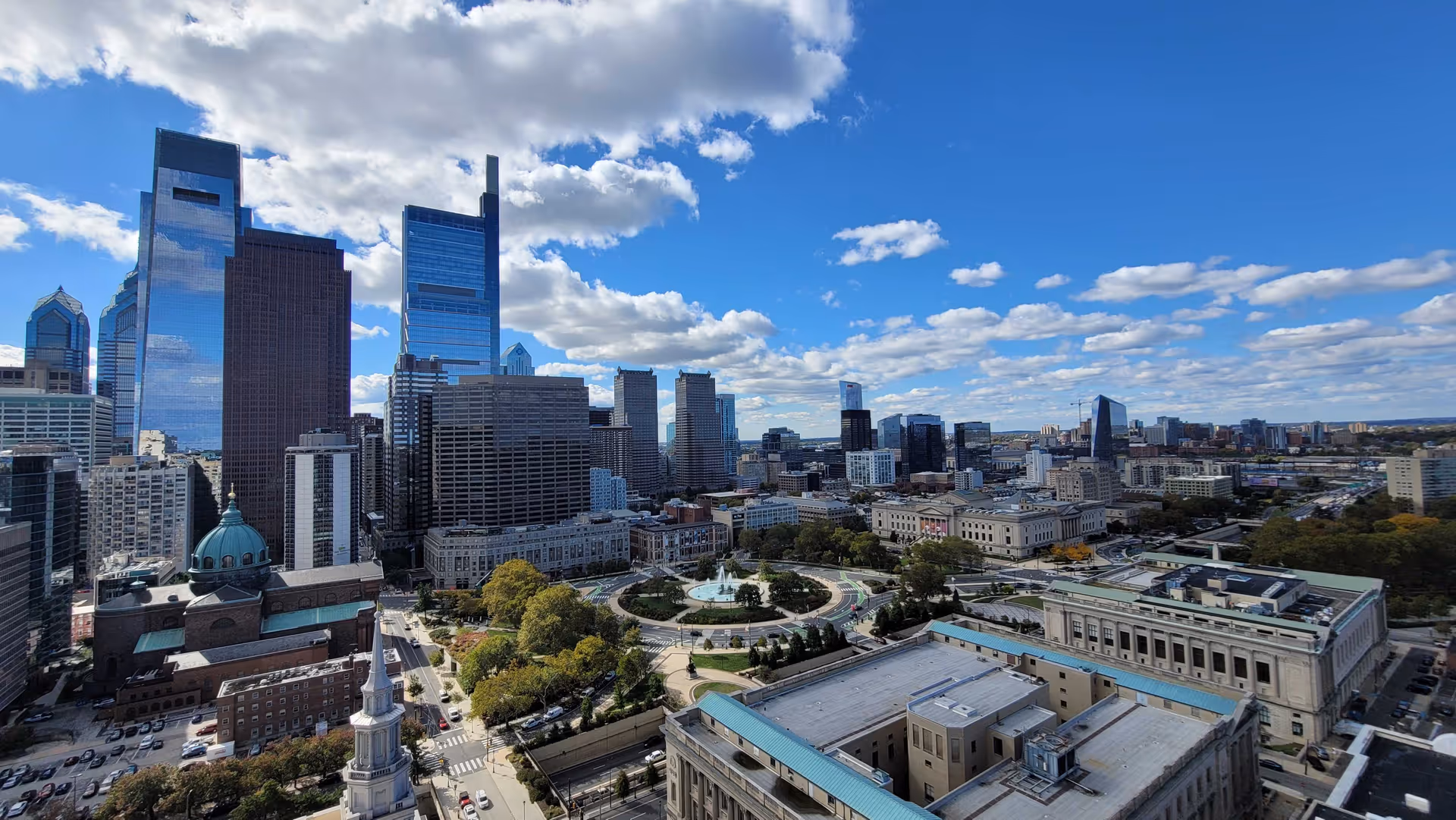Aerial view of a downtown city skyline with skyscrapers, a circular fountain park, and surrounding buildings under a blue sky.
