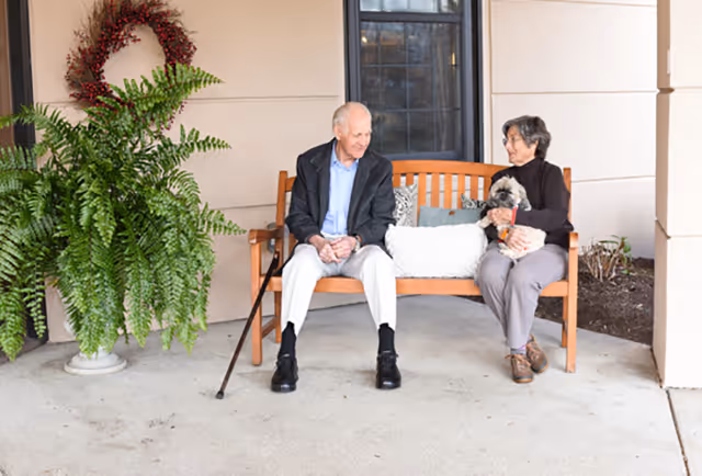 An elderly man and woman sitting on a wooden bench outside a building. The man is holding a cane and the woman is holding a small dog. There is a large potted fern and a decorative wreath on the wall behind them.