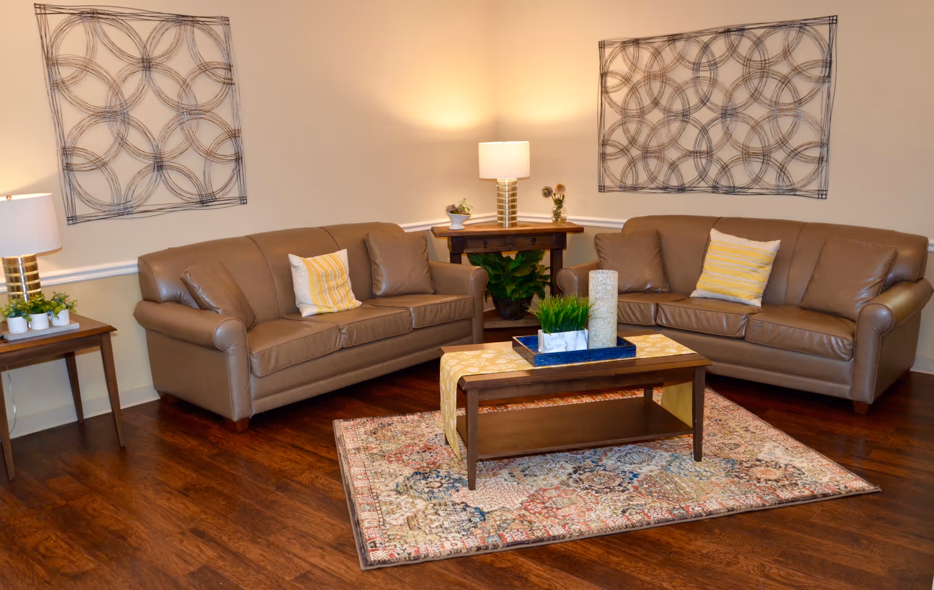 A cozy living room with two brown leather sofas facing each other, each with a yellow and white striped pillow and a brown pillow. Between the sofas is a wooden coffee table with a decorative runner, a tray holding a candle and a small plant. The room has wooden flooring and a patterned area rug. There are two side tables with lamps and plants, and two abstract circular wall art pieces hanging on the beige walls.