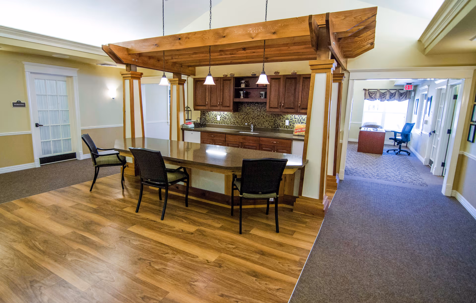 Interior view of a senior living facility kitchen area with wooden cabinetry, a countertop island with three chairs, pendant lights hanging from a wooden beam structure, and a hallway leading to an office space with a desk and chair.
