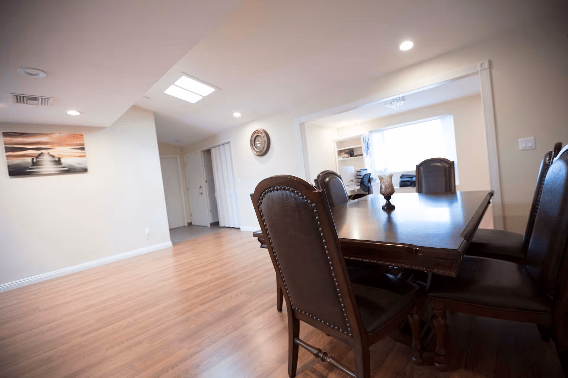 Interior view of a dining area with a dark wooden dining table and six matching chairs with leather upholstery. The room has light-colored walls, wooden flooring, recessed ceiling lights, and a wall clock. There is a large window in the adjacent room letting in natural light, and a decorative painting on one wall.