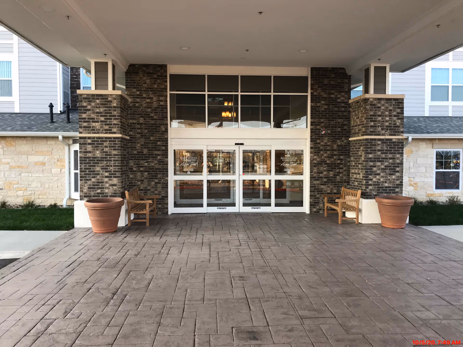 Covered entrance to Aspen Trail Retirement Resort with sliding glass doors flanked by brick columns, benches, and large planters.