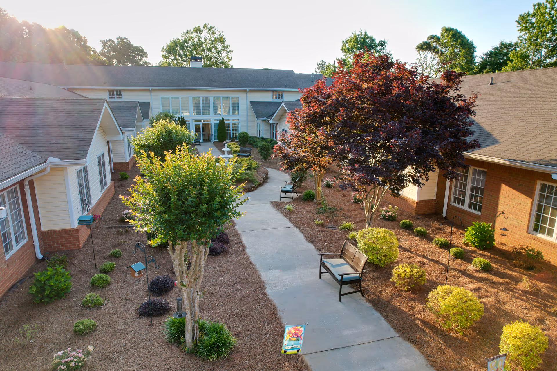Outdoor courtyard area of a senior living facility with a paved walkway, benches, various trees and shrubs, and a fountain in the background. The building surrounds the courtyard with large windows and brick and siding exterior walls. Sunlight filters through the trees creating a warm atmosphere.