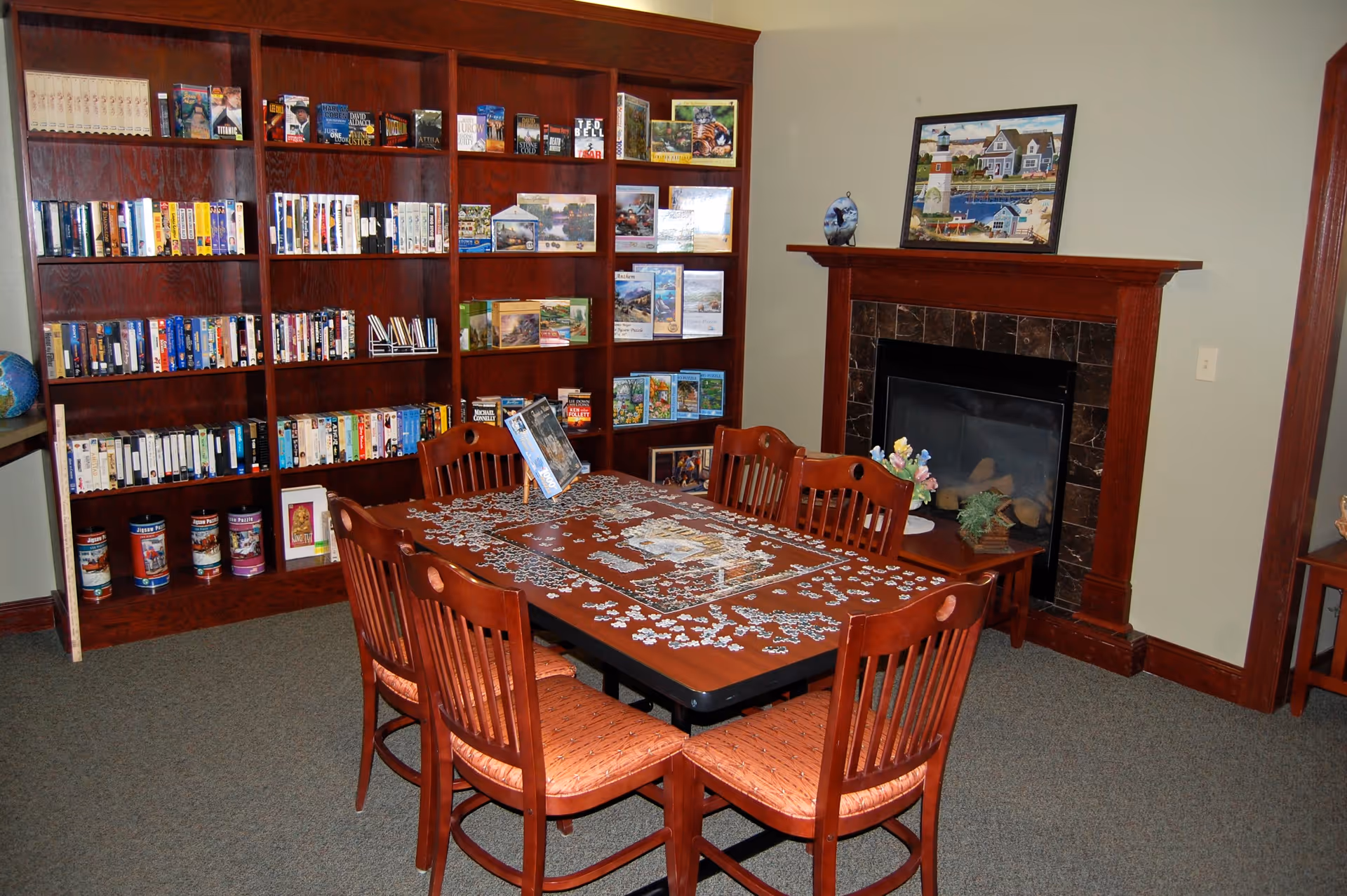 A cozy room with a large wooden table in the center surrounded by six wooden chairs with orange cushions. On the table, there is an incomplete jigsaw puzzle. Behind the table, there is a large wooden bookshelf filled with books and puzzles. To the right, there is a fireplace with a dark tile surround and a wooden mantel, decorated with a framed picture and small decorative items. The room has beige walls and carpeted flooring.