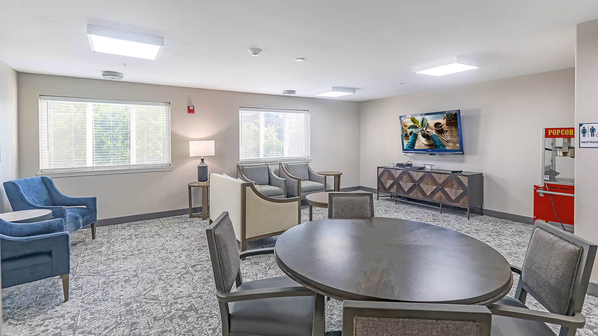 A well-lit common area with a round wooden table surrounded by four chairs in the foreground. In the background, there are two blue armchairs, a beige loveseat, two gray armchairs, and a wooden side table with a lamp. A flat-screen TV is mounted on the wall above a wooden console. A red popcorn machine and restroom signs are visible on the right side of the room. Two large windows with blinds allow natural light to enter.