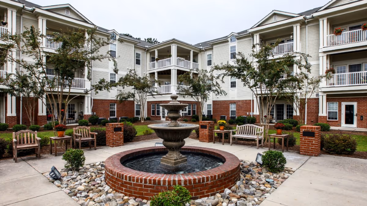 Outdoor courtyard area of McCarthy Court featuring a central brick fountain surrounded by rocks, wooden benches, small tables with potted plants, and trees. The courtyard is enclosed by a three-story building with balconies and windows.