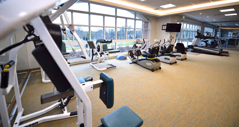 Bright exercise room with cardio and strength machines facing large windows overlooking outdoor grounds.