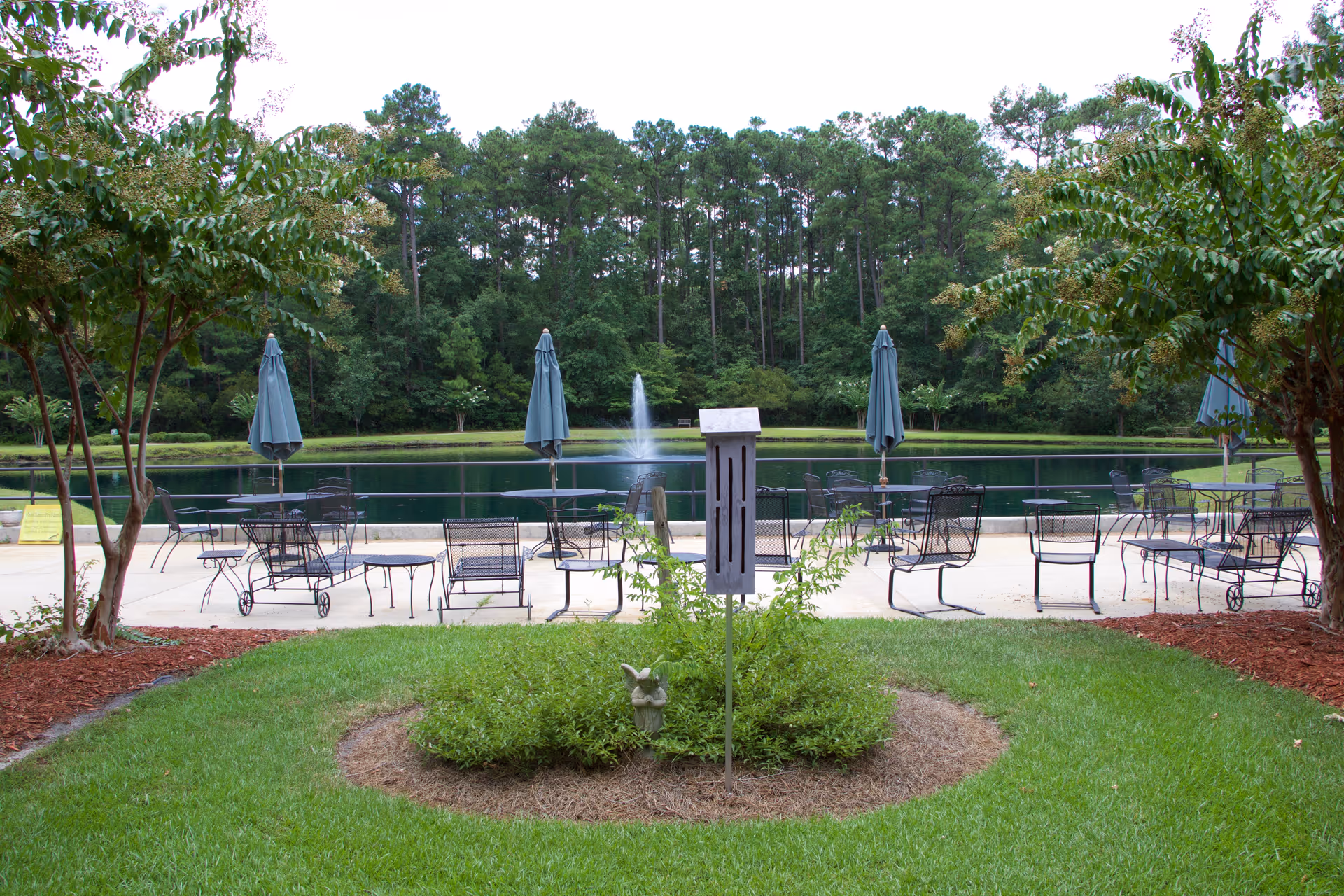 Outdoor patio area with metal tables and chairs, some with closed blue umbrellas, overlooking a pond with a fountain in the center. The area is surrounded by green grass, trees, and landscaped bushes.