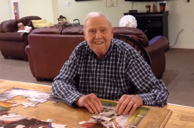 An elderly man wearing a checkered shirt is sitting at a wooden table working on a large jigsaw puzzle. Behind him, there are two brown leather sofas with two elderly individuals seated on them in a cozy living room setting.