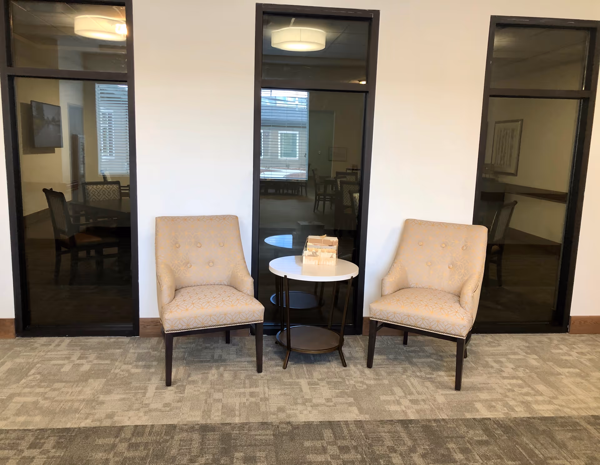 Two beige upholstered chairs with button tufting are placed on either side of a small round white table with a dark metal frame. The table has a wooden block game on top. Behind the chairs are three tall black-framed glass panels showing a dimly lit room with tables and chairs inside. The floor is carpeted with a patterned design.