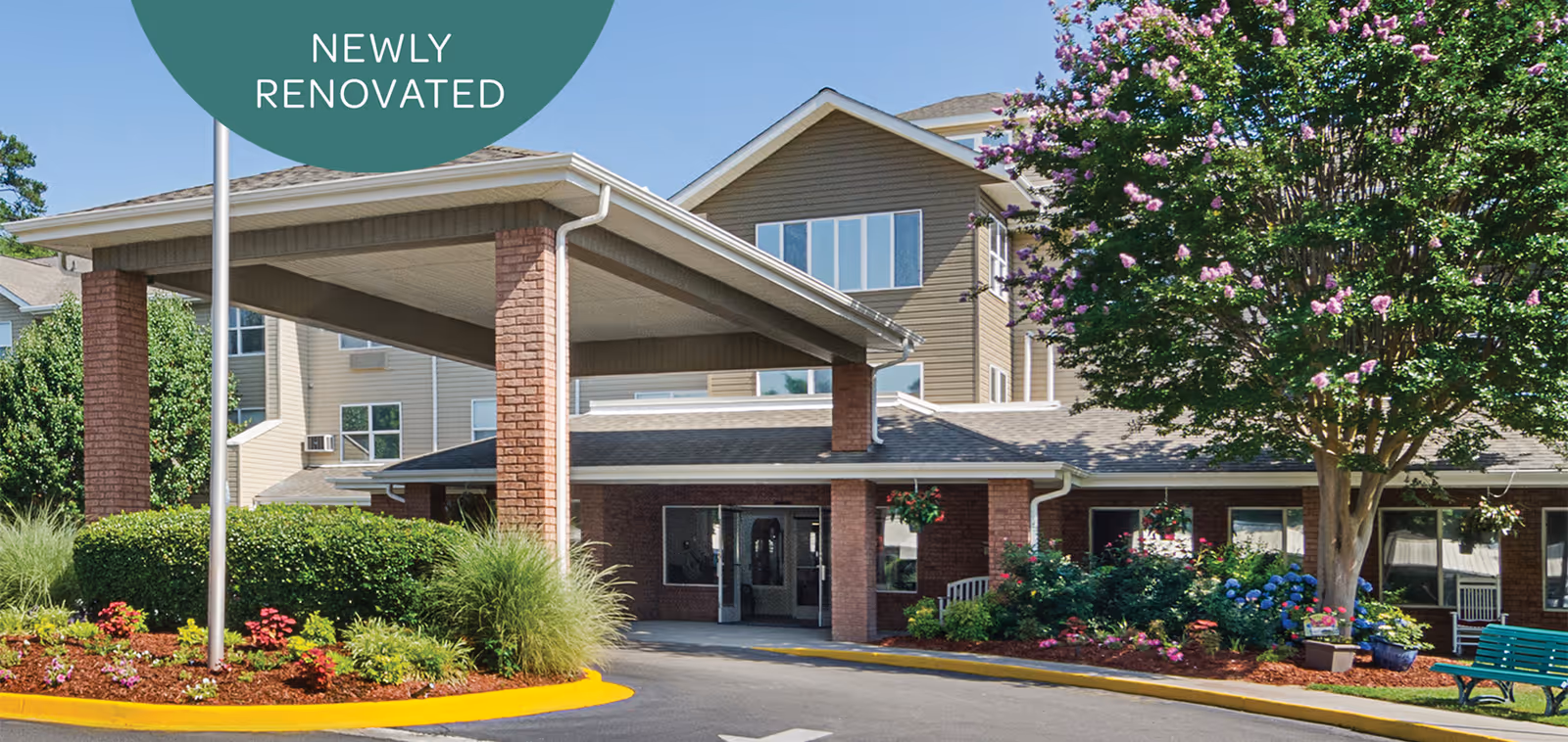 Exterior view of Holiday Columbia Pines senior living facility showing a covered entrance with brick pillars, landscaped garden beds with flowers and shrubs, a large tree with pink blossoms, and a green bench near the entrance. The building has multiple windows and beige siding. A green circular sign above the entrance reads 'Newly Renovated'.