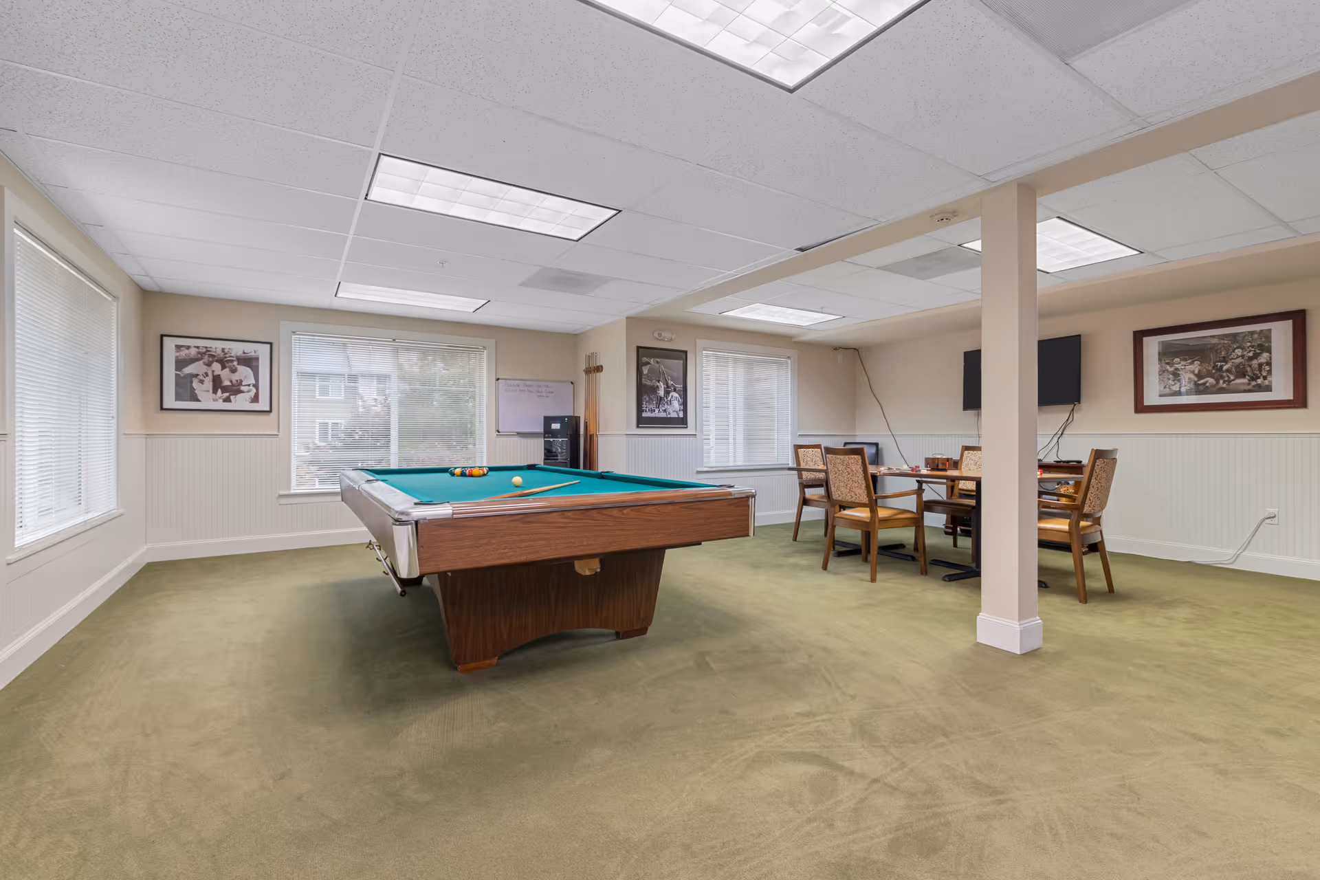 Bright communal game room with a pool table in the center, a table and chairs, wall-mounted TV and framed pictures.