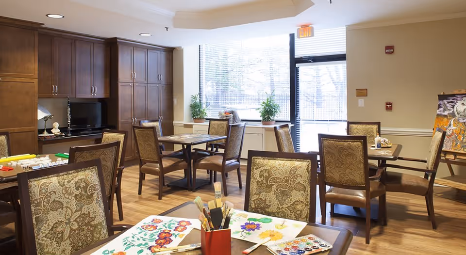 A well-lit activity room with several tables and upholstered chairs. On the tables are art supplies including paintbrushes, watercolor paints, and paper with floral paintings. The room has wooden flooring, large windows with blinds, and built-in wooden cabinets along one wall. There is an exit door with a window and some potted plants on the windowsill.