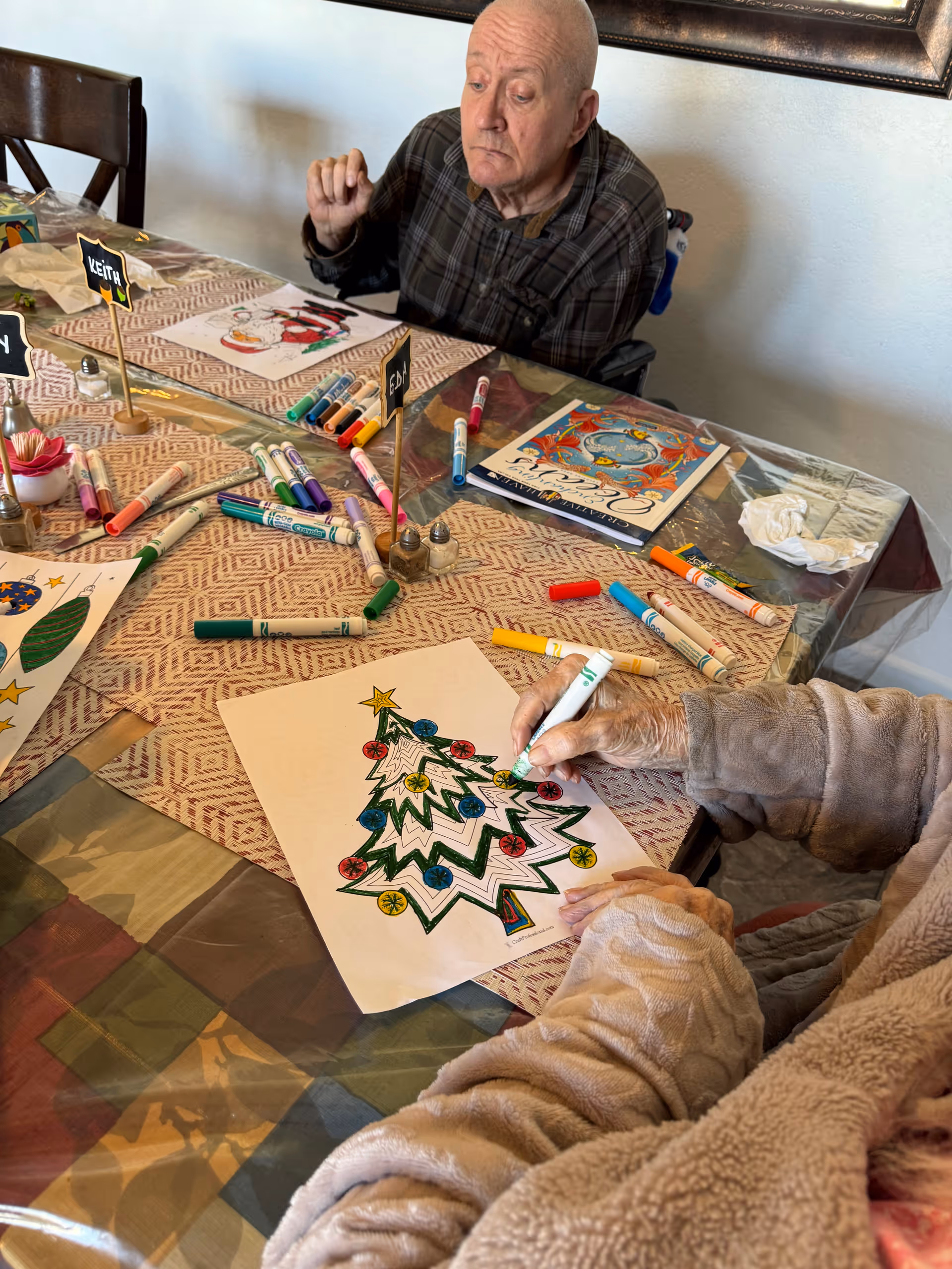 Two elderly individuals seated at a table engaged in coloring holiday-themed pictures. One person is coloring a Christmas tree with markers, while the other looks on. The table is covered with a patterned tablecloth and scattered with various colored markers and coloring sheets.