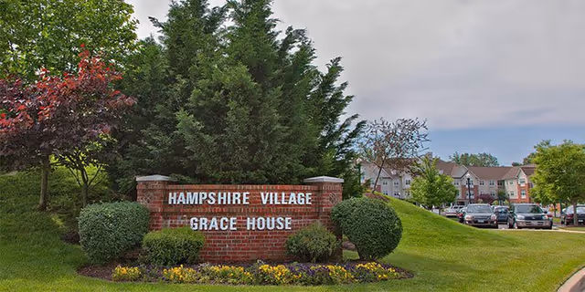 Brick entrance sign reading "Hampshire Village Grace House" on a landscaped lawn with trees and the facility building and parked cars in the background.