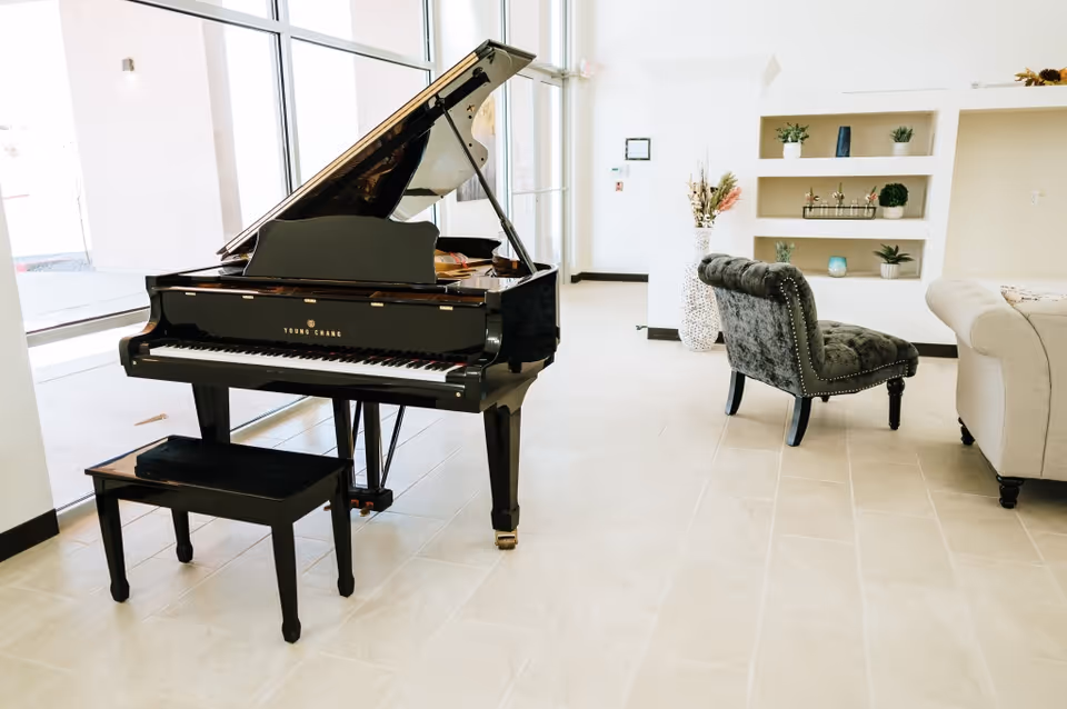Bright and spacious room with a black grand piano and matching bench near large windows. The room features light-colored tiled flooring, a gray upholstered chair with studded detailing, a beige sofa, and built-in shelves decorated with small plants and ornaments.