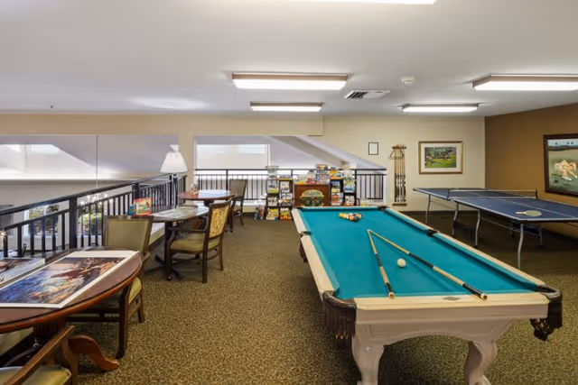 A senior community game room with a pool table and cues in the foreground, a ping-pong table to the right, and card tables, chairs and shelves of board games in the background.