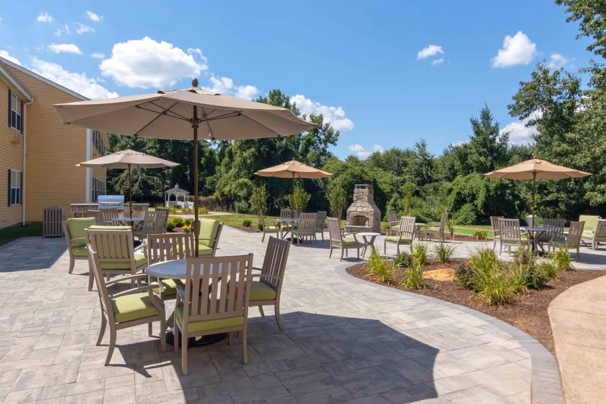 Outdoor patio area with multiple tables and chairs, each shaded by large beige umbrellas. The patio is paved with stone tiles and surrounded by landscaped greenery and trees. A stone fireplace is visible in the background under a clear blue sky with scattered clouds.