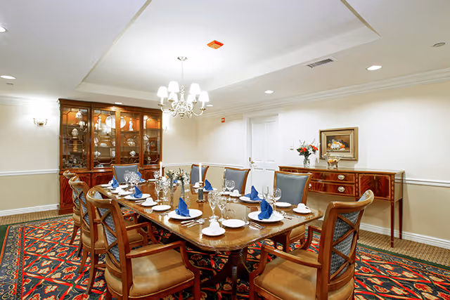A formal dining room with a long wooden table set for a meal, featuring blue folded napkins, white plates, and glassware. The room has wooden chairs with cushioned seats, a chandelier hanging from the ceiling, a patterned carpet, a wooden china cabinet displaying dishes, and a sideboard with a vase of flowers and a framed picture on the wall.