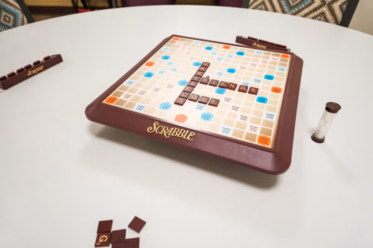 A Scrabble board game set up on a white round table with letter tiles forming the words GARFIELD, FRIENDS, and LOVE. There are Scrabble tile racks and a sand timer on the table, with patterned chairs in the background.