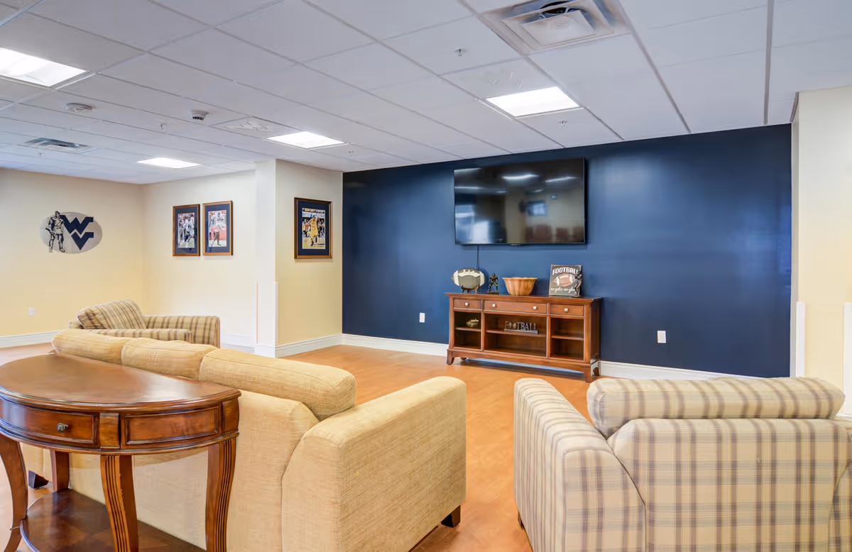 A cozy living room area with beige and plaid upholstered sofas and armchairs arranged around a wooden console table and a large flat-screen TV mounted on a dark blue accent wall. The room has light-colored walls, wood flooring, and framed sports-themed pictures on the wall.