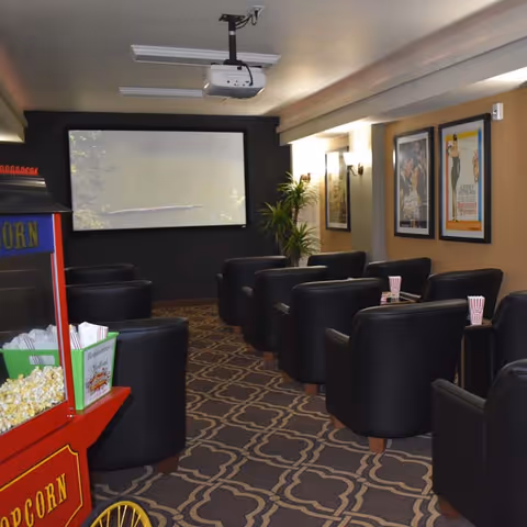 A cozy movie theater room with black leather armchairs arranged in rows facing a large projection screen. There is a popcorn machine on the left side and framed movie posters on the right wall. The room has patterned carpet and a ceiling-mounted projector.