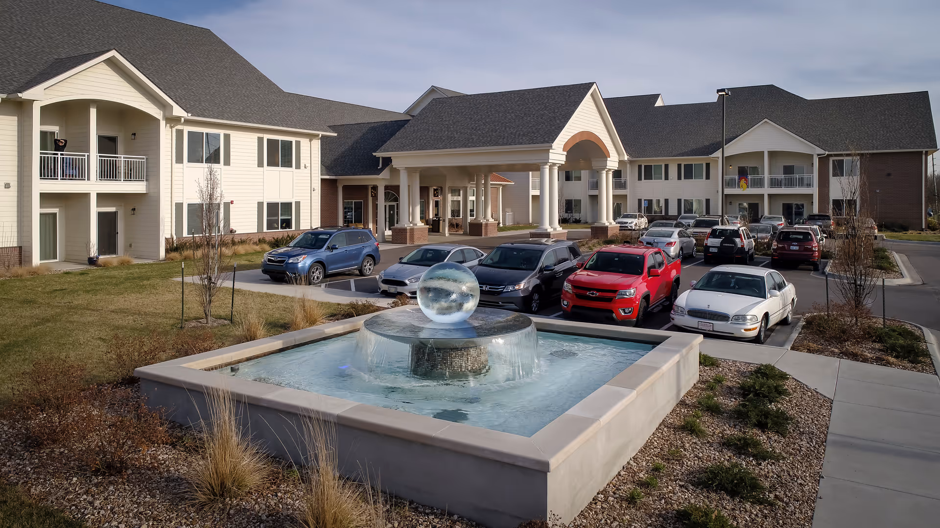Exterior view of Pioneer Ridge Independent Living of Lawrence showing a large building with multiple windows and balconies, a covered entrance with columns, a parking lot with several cars, and a decorative water fountain in the foreground surrounded by landscaping.
