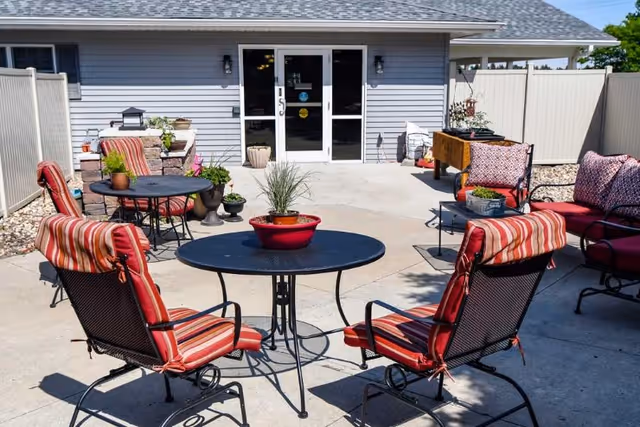 Outdoor patio area with metal tables and chairs featuring red and striped cushions. There are potted plants on the tables and around the patio. The patio is enclosed by a white fence and is adjacent to a building with double glass doors.