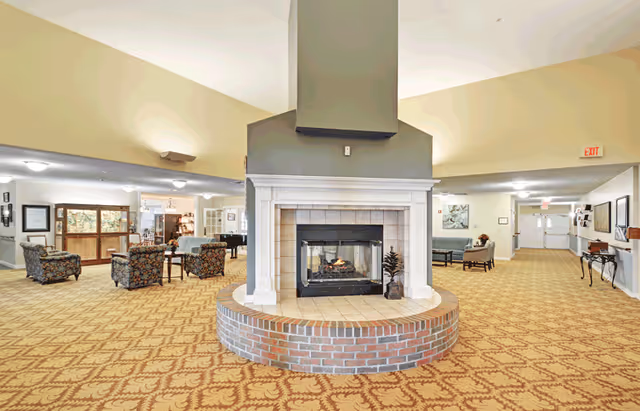 Spacious senior living facility common area with a central double-sided fireplace surrounded by a brick base. The room features patterned carpet, multiple seating areas with floral upholstered chairs and sofas, and bright lighting. There are exit signs and framed artwork on the walls.