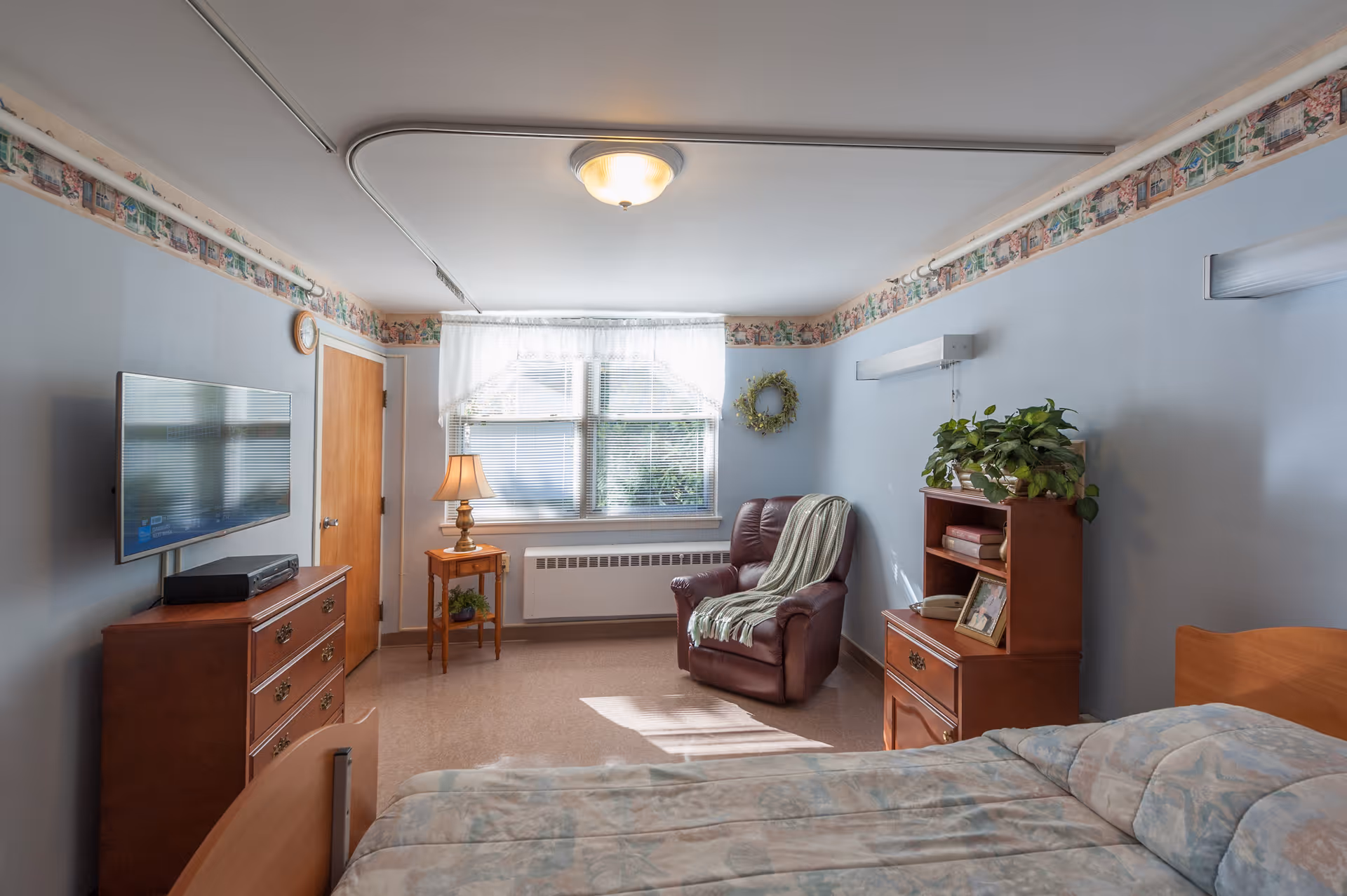 A cozy senior living bedroom with a bed in the foreground, a wooden dresser with a flat-screen TV mounted above it on the left wall, a small wooden side table with a lamp next to a window with sheer curtains, and a comfortable leather armchair with a throw blanket. There is also a wooden nightstand with a plant, books, a phone, and a framed photo. The walls are painted light blue with a decorative border near the ceiling.
