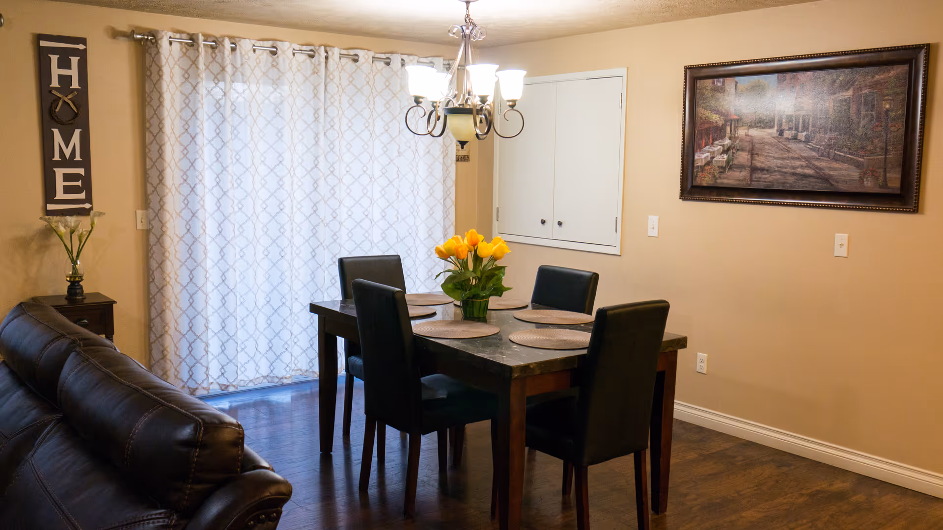 A dining area with a dark wooden table set for four with round placemats and a vase of yellow tulips in the center. Four black chairs surround the table. To the left, part of a dark brown leather couch is visible. A wall decoration with the word 'HOME' hangs on the left wall above a small table with a vase of flowers. A large window with patterned curtains is in the background. On the right wall, there is a framed painting and a white cabinet with double doors. A chandelier with five lights hangs above the dining table.