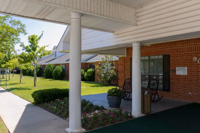 Covered entrance of a brick senior living building with white columns, rocking chairs, flowerbeds and a sidewalk beside a green lawn.
