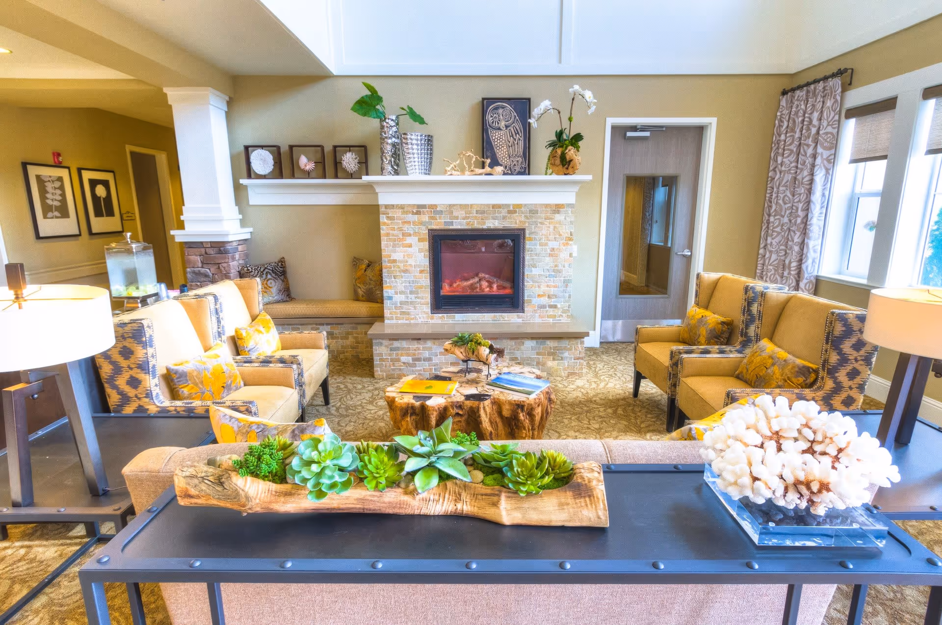 A cozy living room in Carmel Village Memory Care featuring a stone fireplace with decorative items on the mantel, comfortable armchairs with patterned cushions arranged around a wooden coffee table, a sofa with a planter of succulents on a console table behind it, and large windows with patterned curtains allowing natural light to fill the space.
