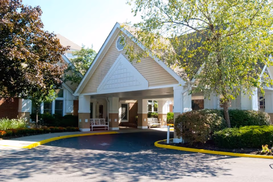 Covered entrance canopy and circular driveway at a senior living facility with landscaped shrubs and trees.