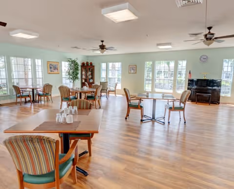 A spacious dining room with several wooden tables and chairs arranged neatly. The room has large windows allowing natural light to fill the space, light green walls, ceiling fans, and a piano in the corner. There are also some decorative plants and framed pictures on the walls.