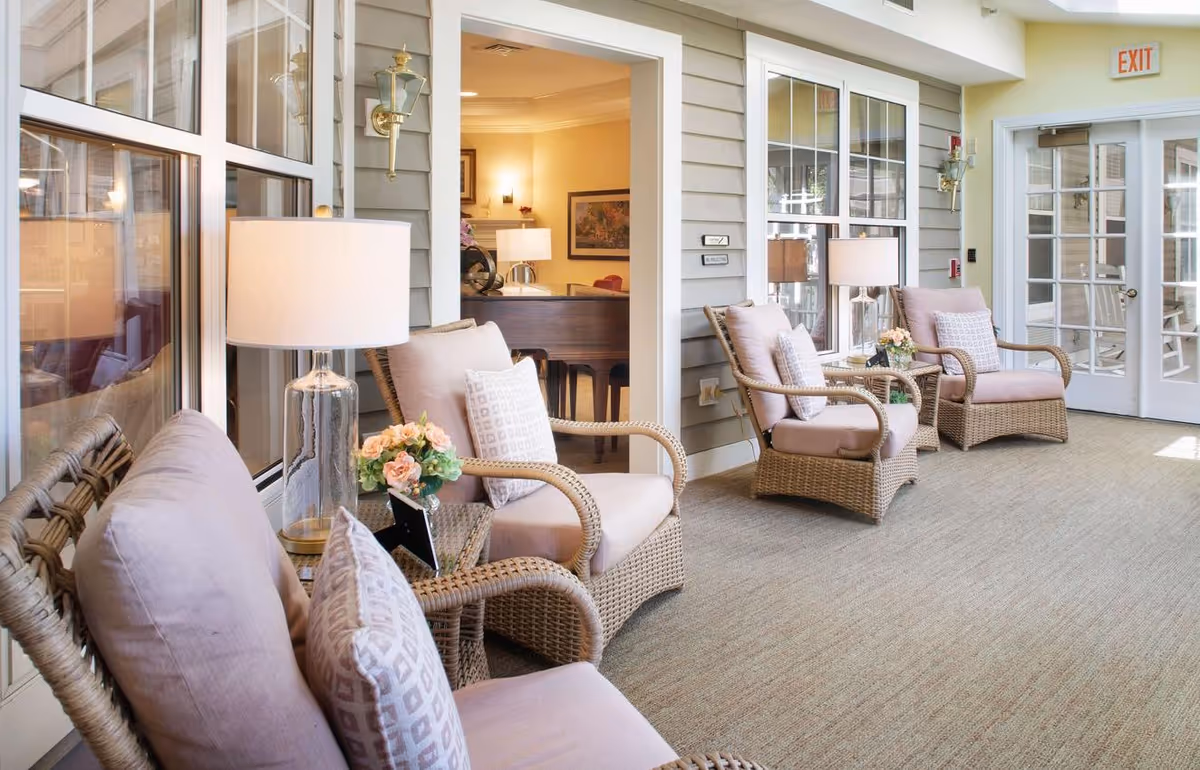 Bright indoor sitting area with wicker armchairs, side tables, lamps, and floral arrangements along windows and glass doors.