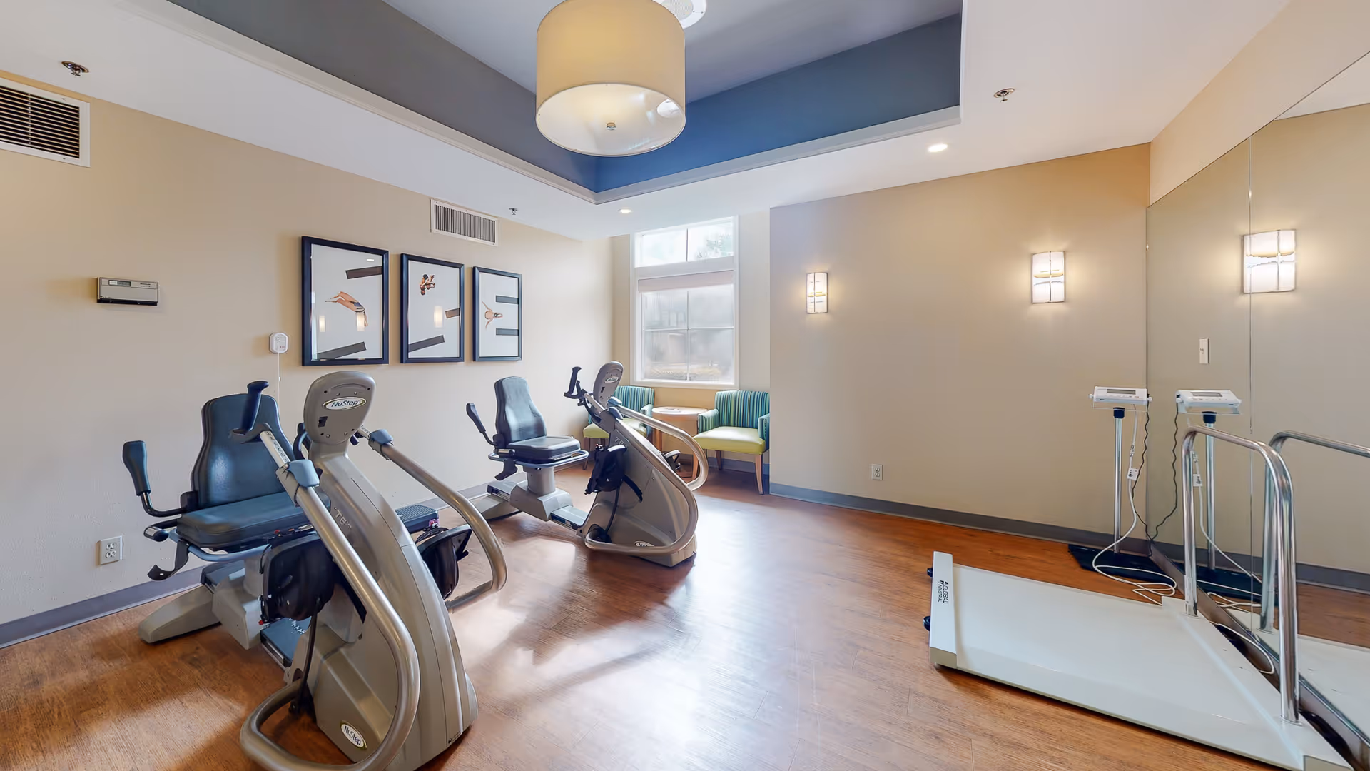 A small fitness room with two recumbent exercise bikes, a white treadmill, and two green striped chairs near a window. The room has wood flooring, beige walls, a large mirror on one side, and a modern ceiling light fixture.