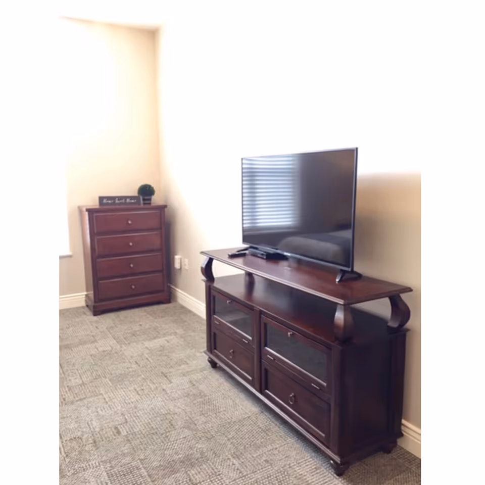 A corner of a room with a wooden TV stand holding a flat-screen television and a wooden chest of drawers with a small potted plant and a decorative sign on top. The room has light-colored walls and carpeted flooring.